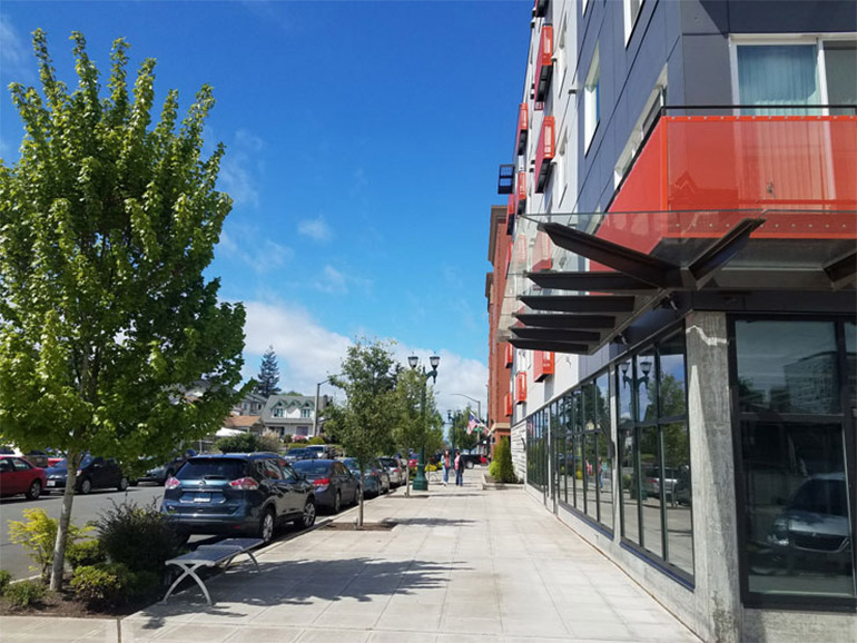 A sidewalk separates an apartment building and a line of parked cars along a street.